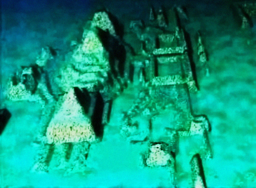 Underwater structures resembling ancient stone pyramids and rectangular formations, partially covered by sand, illuminated by blue-green light, suggesting possible archaeological ruins on the ocean floor.