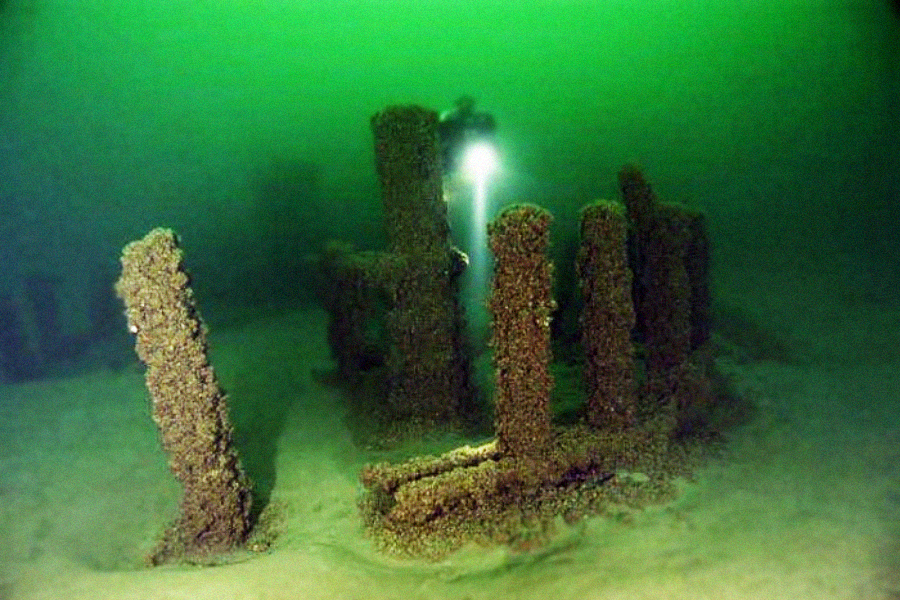 A scuba diver with a flashlight explores an underwater shipwreck covered in algae and marine growth, surrounded by green-tinged water and sandy seabed.