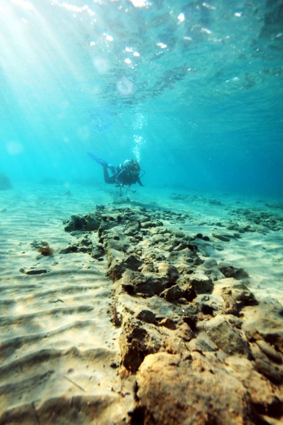 A scuba diver swims above a rocky path on the sandy ocean floor, with sunlight filtering through clear blue water from above.