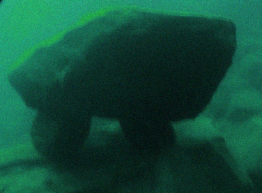 A large, irregularly shaped rock formation underwater, with a greenish tint and two smaller rocks supporting it, creating a table-like appearance on the seafloor.