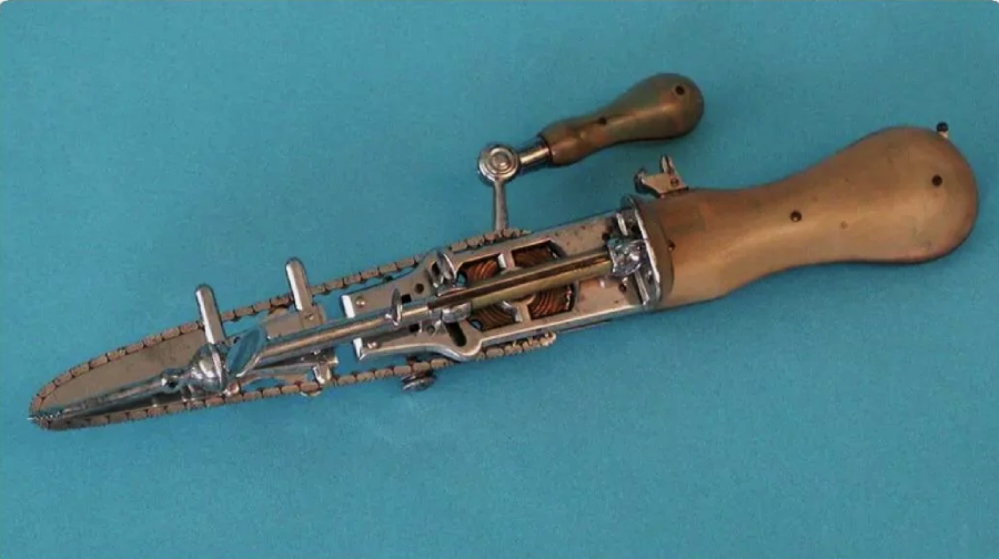 A vintage medical tool with a wooden handle and a small chain saw blade, set against a blue background. The device resembles a miniature chainsaw and was historically used for surgical procedures.