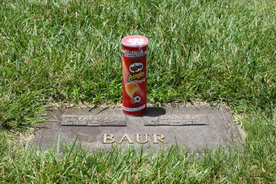 A can of Pringles Original chips is placed on a grave marker that reads "Fred J. Baur" amidst green grass.