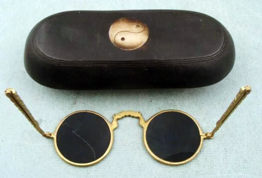 A pair of vintage round sunglasses with gold frames and black lenses sits below a black eyeglass case featuring a yin-yang symbol. The background is a light, textured surface.