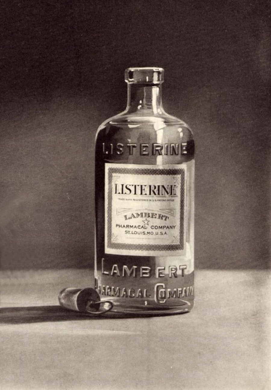 A vintage glass bottle of Listerine mouthwash with embossed lettering and a label reading "Lambert Pharmacal Company, St. Louis, MO, USA" is shown next to a bottle cap on a flat surface.