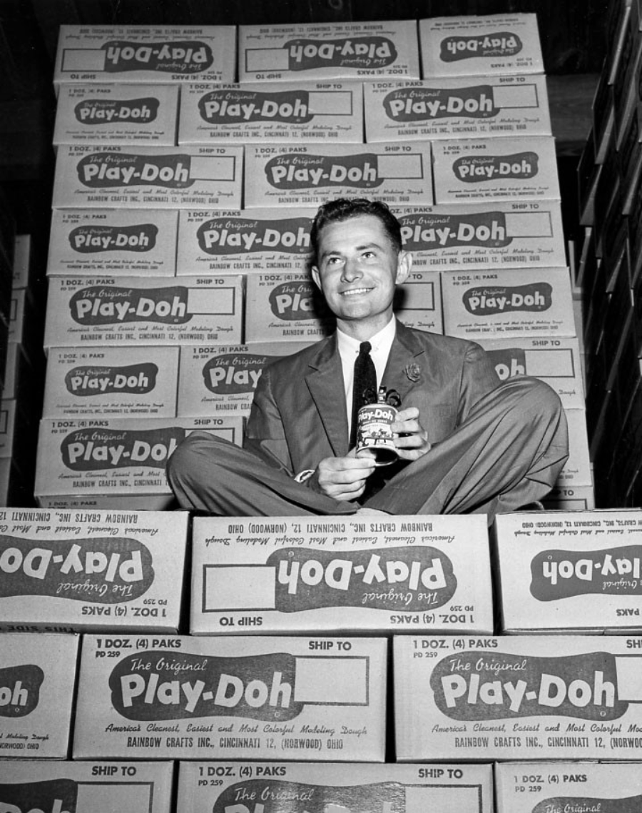 A smiling man in a suit sits cross-legged atop stacked boxes of Play-Doh, holding a Play-Doh can, surrounded by more boxes marked “Play-Doh” filling the background.