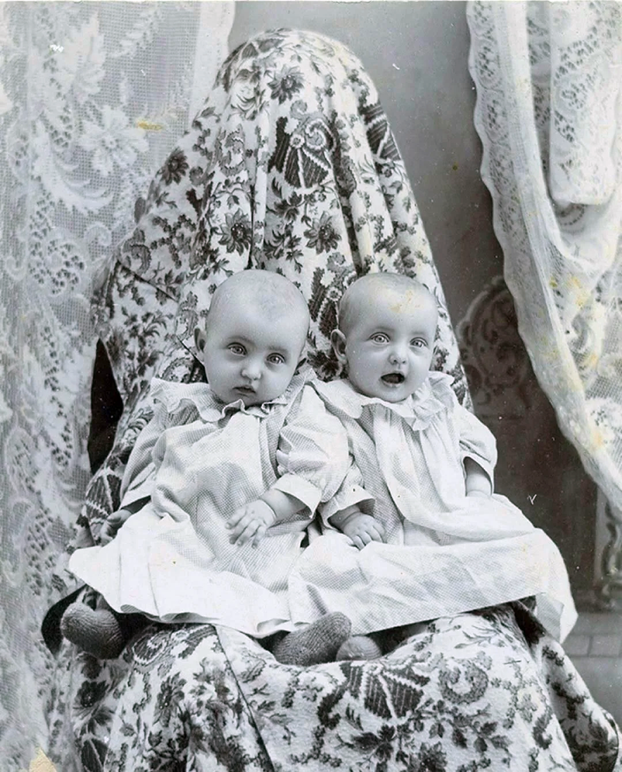 Two babies in old-fashioned dresses sit on a patterned chair. Behind them, an adult figure, covered entirely with a patterned cloth, is mostly concealed, blending into the background curtains.
