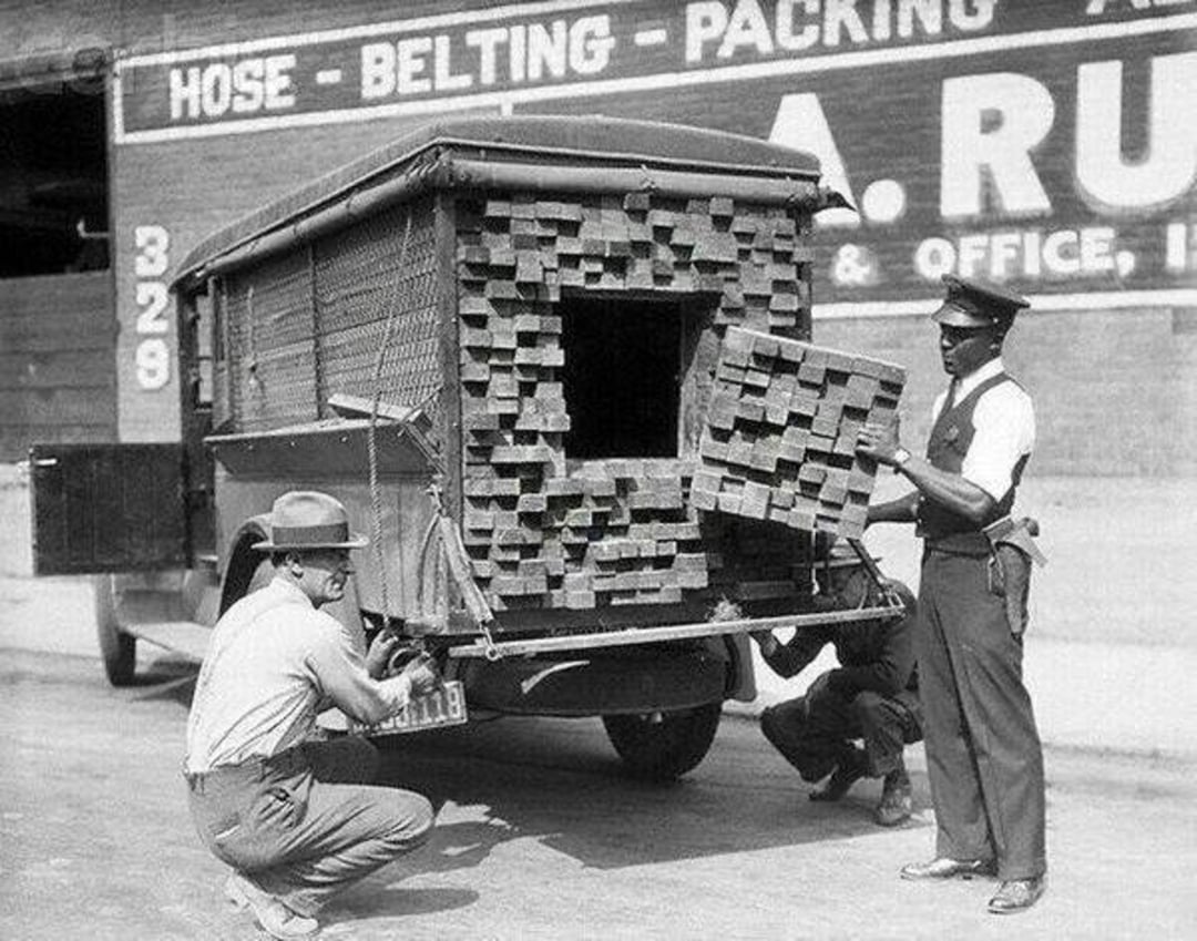 Two policemen inspect the back of a truck filled with bricks, revealing a hidden compartment. One officer holds a brick, while another examines the vehicle. A warehouse sign is visible in the background.