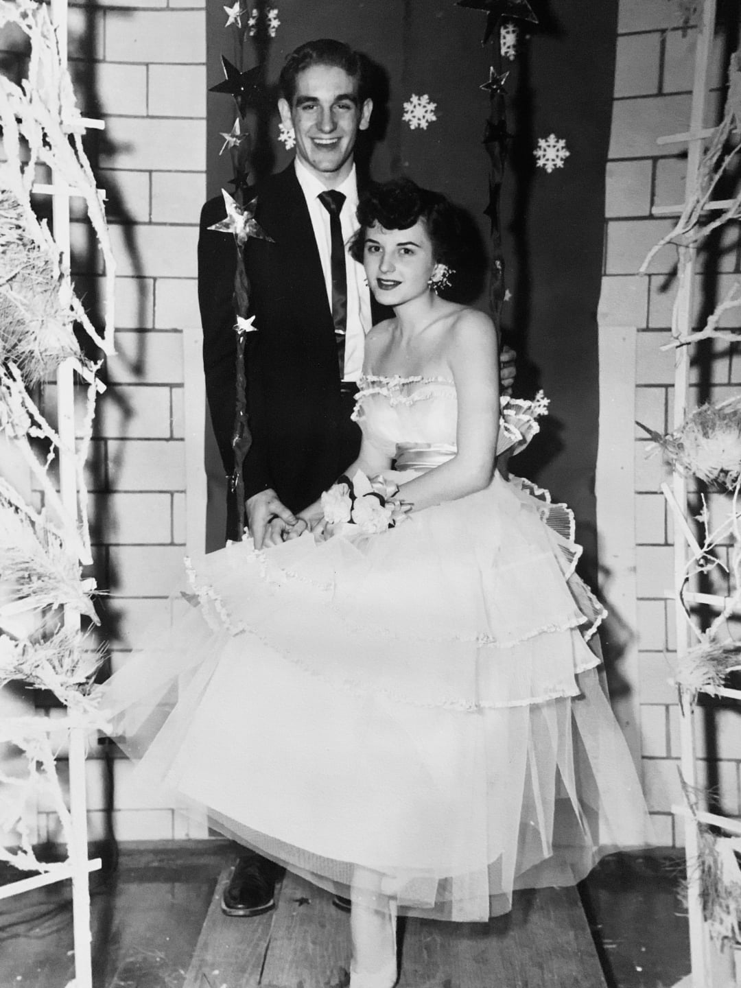 A young couple dressed in formal attire poses together at a dance or prom, smiling. The woman wears a layered, strapless tulle dress and corsage, while the man is in a dark suit and tie. Snowflake decorations hang in the background.
