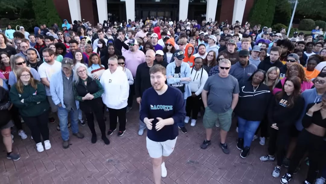A man in a blue shirt and shorts stands at the center of a large crowd of diverse people gathered outdoors on a paved area in front of a building, all looking toward the camera.
