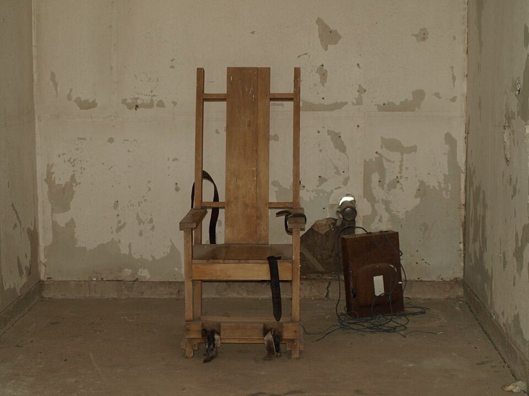 A worn wooden electric chair with leather restraints stands in the center of a bare, peeling, and dimly lit room. Exposed wires and an old electrical box are placed on the floor beside the chair.