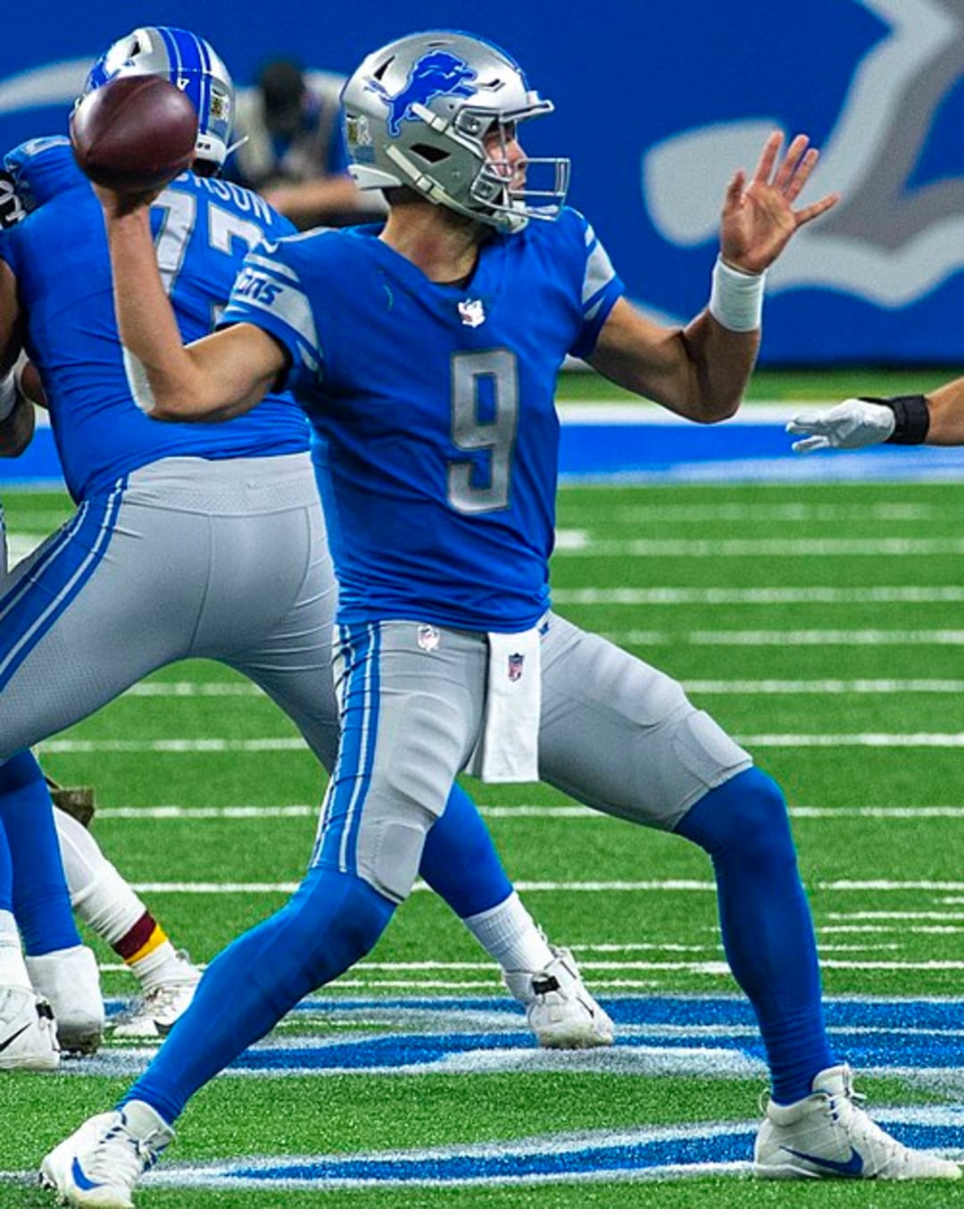 A Detroit Lions football player in a blue and silver uniform prepares to throw a pass during a game on a football field.