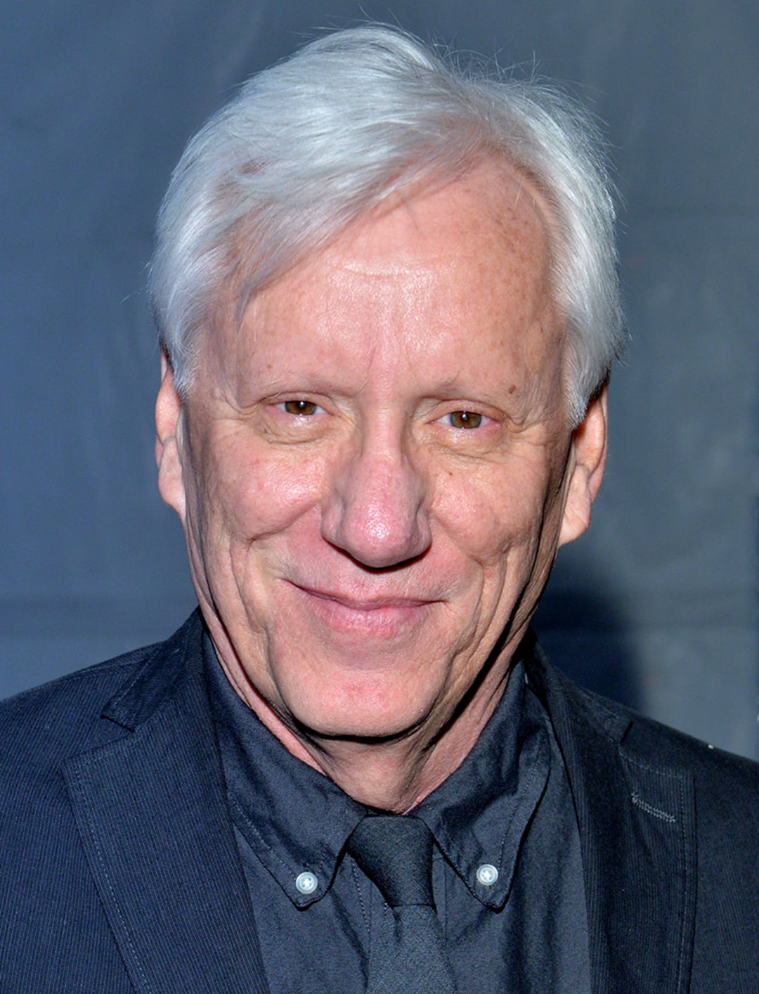 A smiling older man with short white hair wearing a dark suit, shirt, and tie, standing in front of a plain grey background.