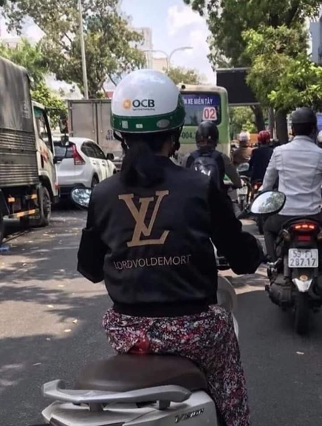 A person riding a scooter in traffic wears a black jacket with a gold "LV" logo and the words "LORD VOLDEMORT" beneath it. Other riders and vehicles are visible on the busy street.