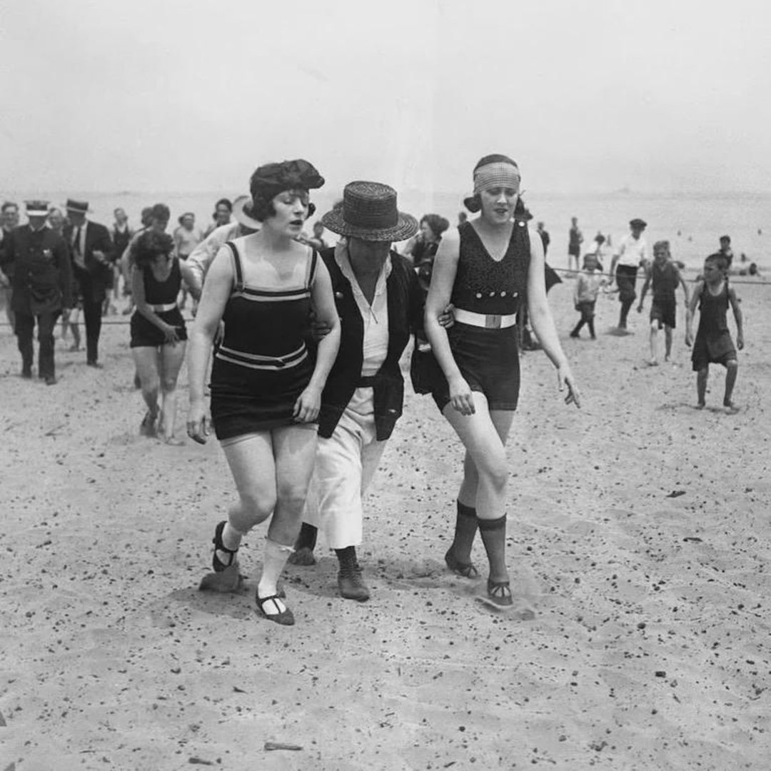 Two women in early 20th-century swimsuits walk on a crowded beach, accompanied by a man in a hat and jacket. Other people, including children, are visible in the background enjoying the beach.