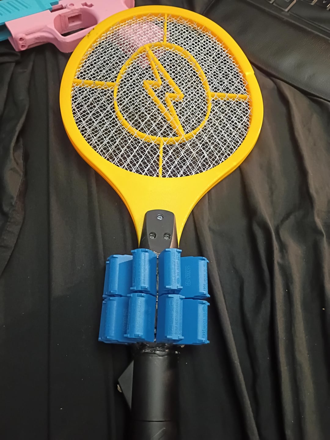 A yellow electric fly swatter with a black handle is lying on a black surface. The handle has blue cylindrical attachments, and part of a pink object is visible in the corner.