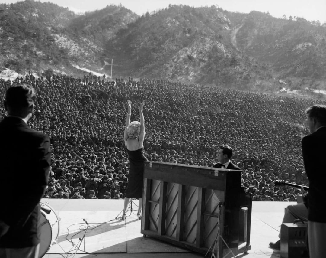 A woman performer with arms raised faces a massive outdoor crowd, backed by musicians and a piano, with mountains in the background. The audience stretches far into the distance.