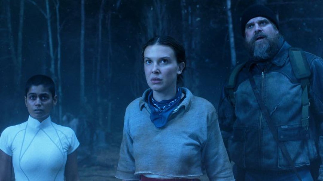 Three people stand in a dimly lit forest, looking ahead with serious, tense expressions. The scene has a blue, eerie glow. One person wears a white outfit, another a scarf and t-shirt, and the third a beanie and jacket.
