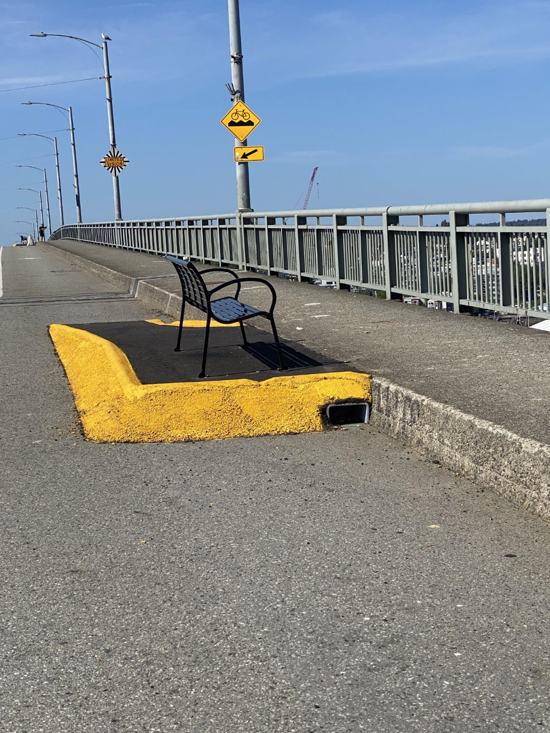 A black metal bench is placed awkwardly on a narrow, yellow-painted raised island beside a road on a bridge, making it unusable. Street signs and railings are visible under a clear blue sky.