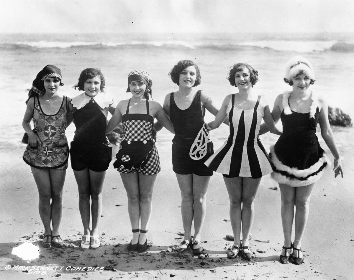 Six women in vintage 1920s swimsuits stand arm in arm on a beach, smiling at the camera with the ocean and waves in the background.