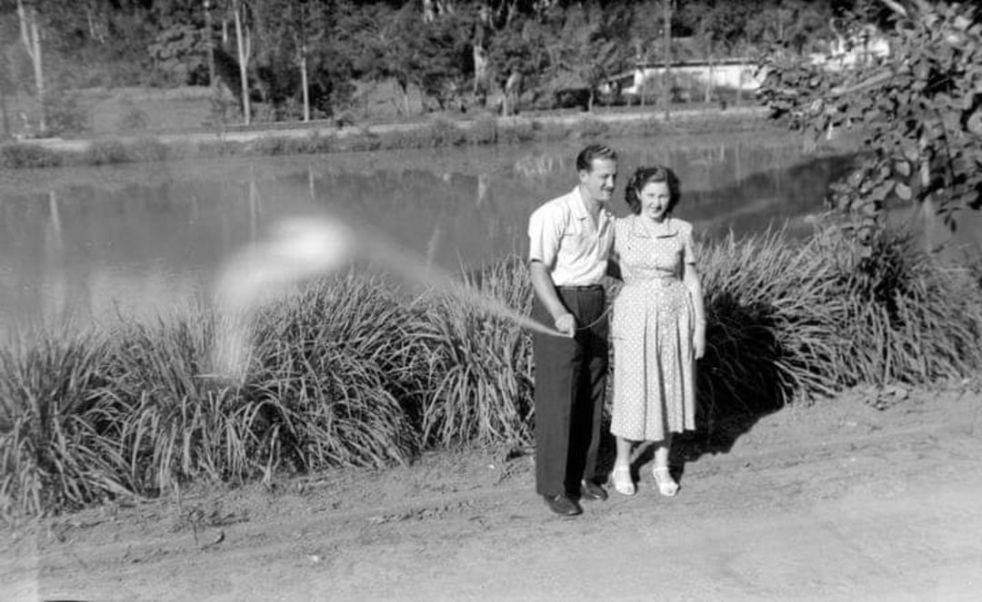 A black-and-white photo shows a smiling man and woman standing by a pond. A blurred, white, rod-like object appears in the foreground, creating an unusual visual effect between them. Trees and shrubs are in the background.