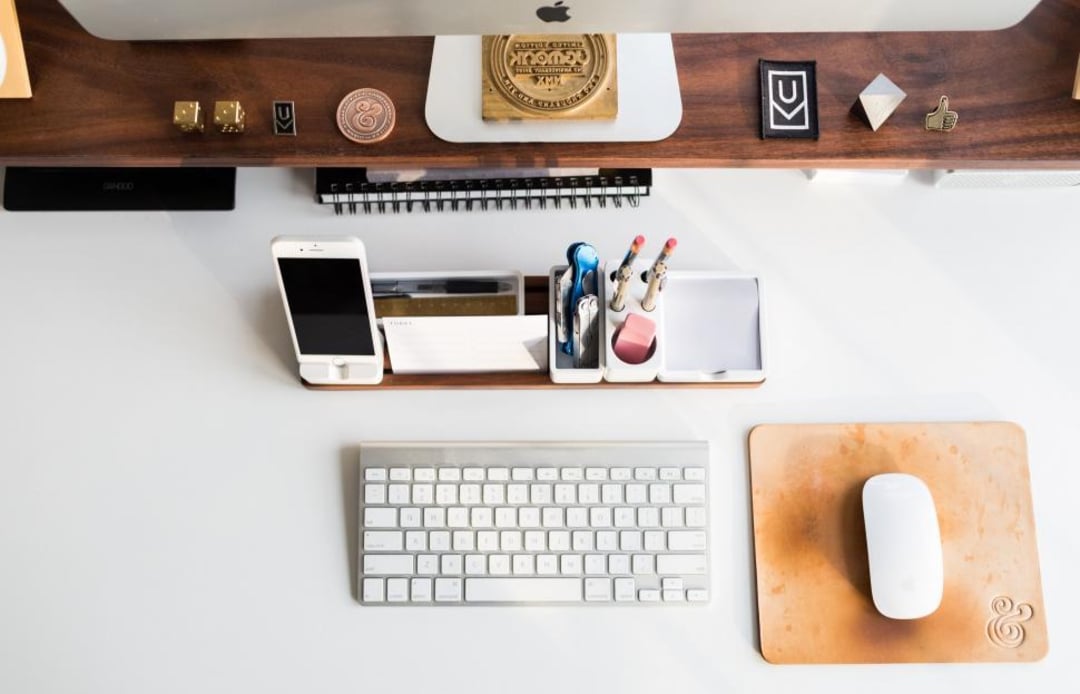 A tidy desk with a computer monitor, wireless keyboard, white mouse on a brown mousepad, and an organizer holding pens, pencils, a phone, and office supplies on a clean white surface.