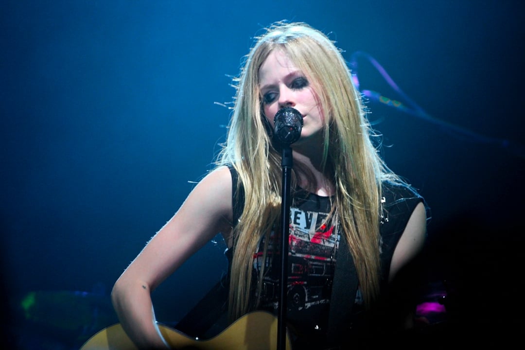A blonde woman sings into a microphone on stage under blue lighting, wearing a sleeveless black graphic shirt and holding a guitar.