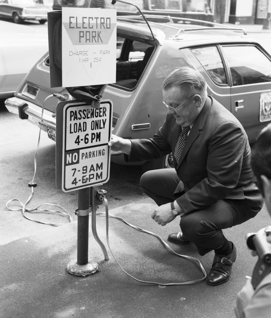A man in a suit kneels beside an electric car, plugging it into a street charging station labeled "Electro Park." Parking signs and cables are visible, and a passerby with a camera stands nearby.