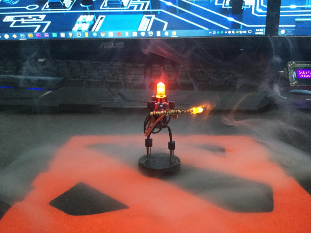 A small robot model with glowing red and yellow lights stands on a desk surrounded by swirling smoke, with a computer keyboard and monitor in the background.