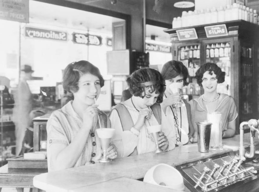 Four young women in 1920s attire sit at a soda fountain counter, smiling and drinking milkshakes with straws. The background shows shelves, glassware, and a window with signage.