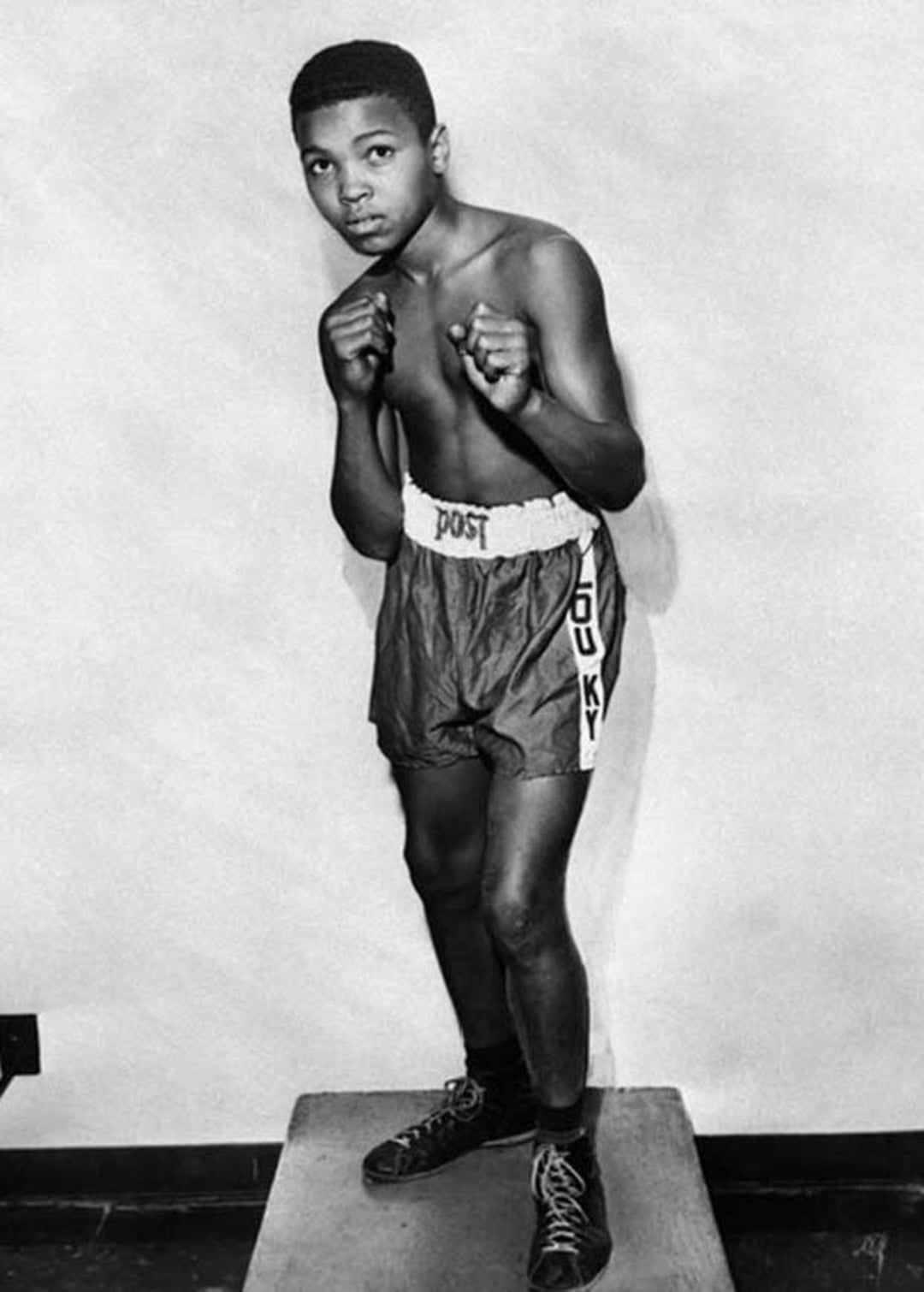 A young boy in boxing shorts and shoes stands on a small platform in a boxing stance, fists raised and facing forward against a plain background.