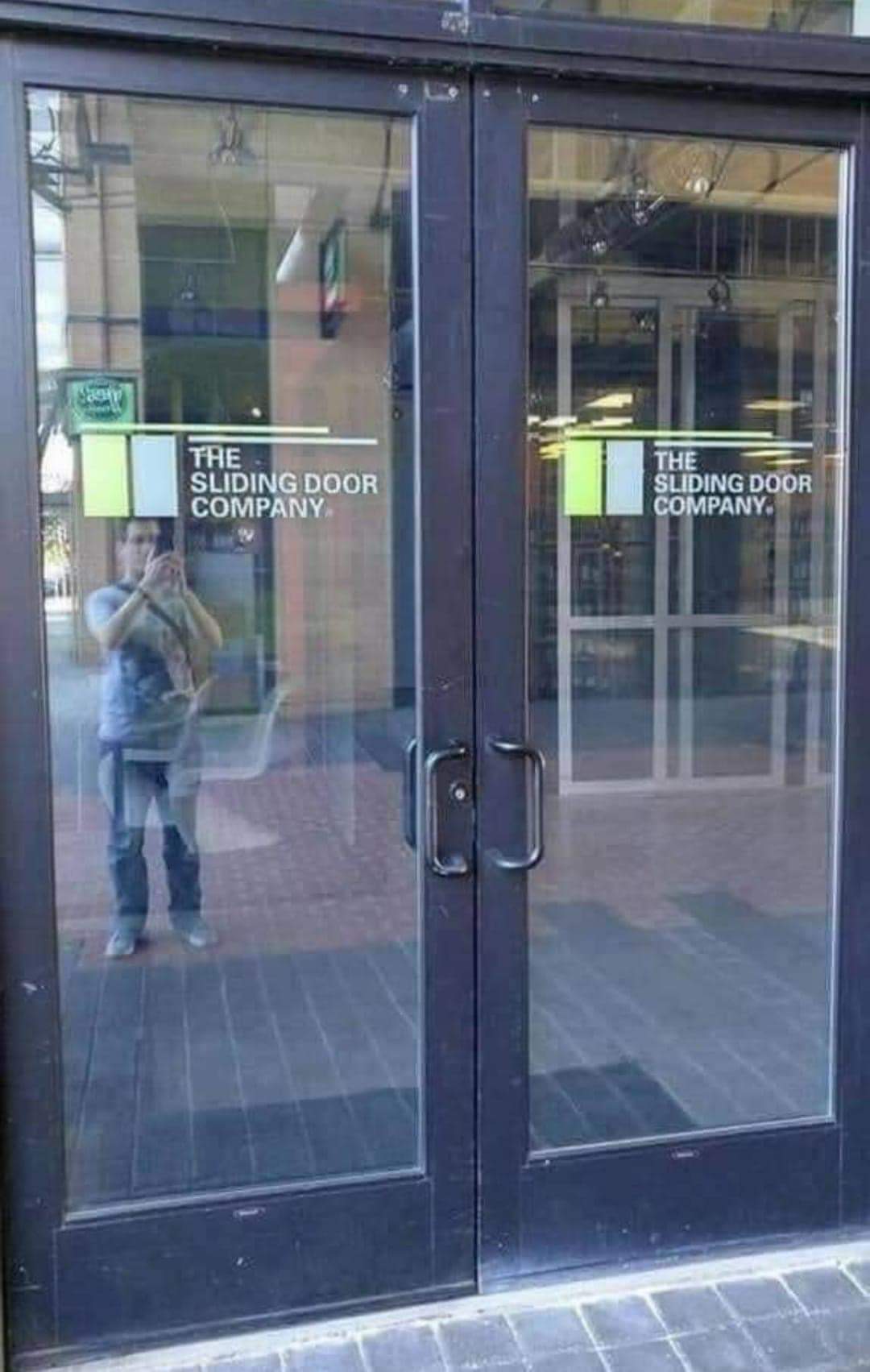 Glass double doors with handles, labeled "The Sliding Door Company." The doors have reflections of a man taking a photo, and the interior of a building is visible through the glass. The doors are not sliding.