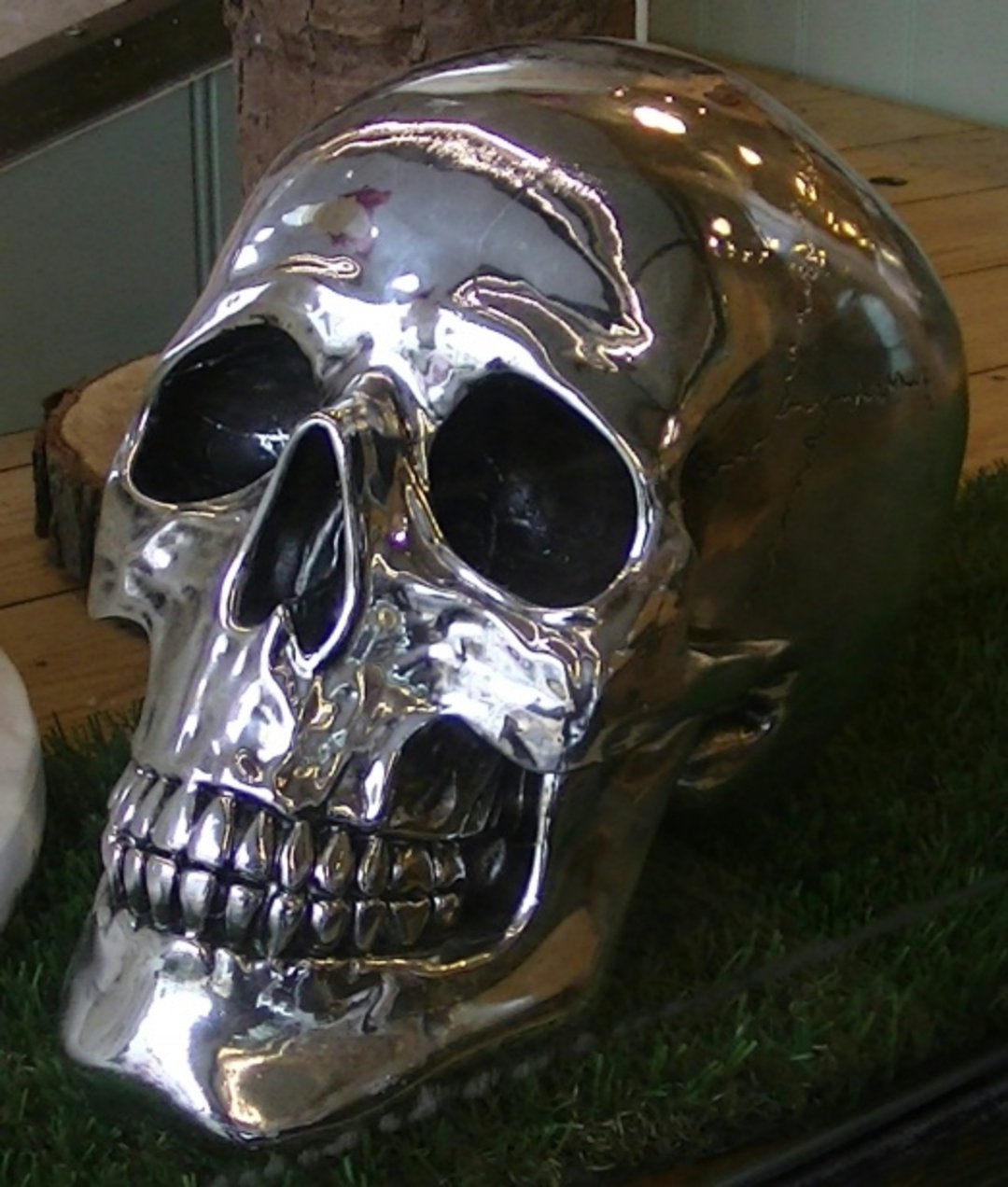A shiny metallic skull sculpture with reflective surfaces rests on a patch of artificial grass, with a wooden floor and part of a tree trunk visible in the background.