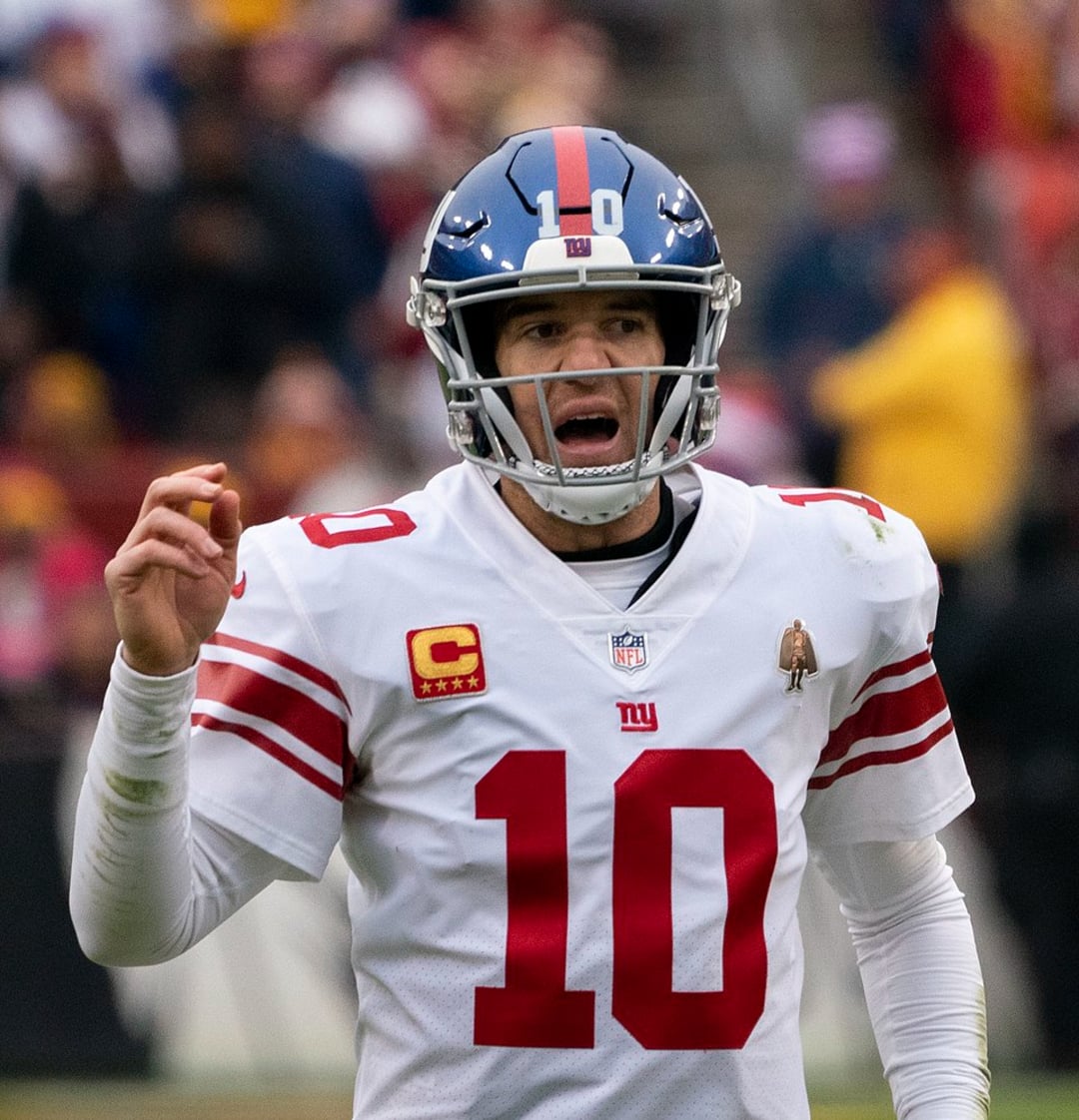 A football player in a white New York Giants uniform and helmet, number 10, stands on the field with his mouth open and hand raised, appearing to call out a play during a game.