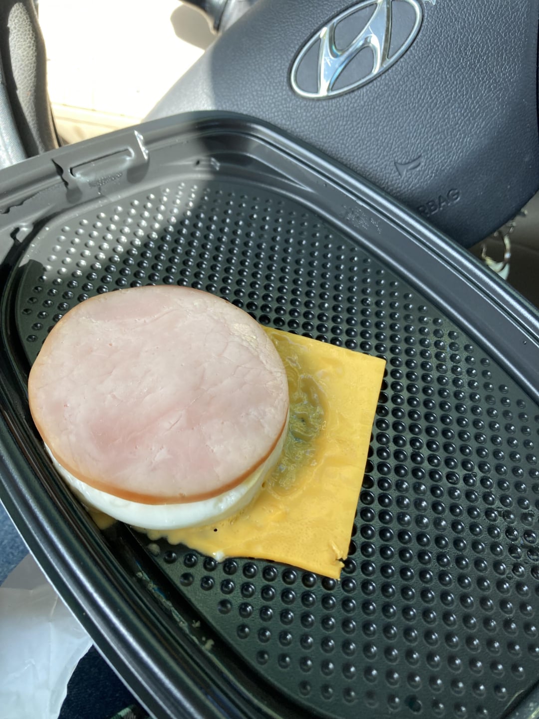 A slice of ham, a fried egg, and a slice of cheddar cheese are in a black plastic takeout container inside a car. The cheese is partly melted and not fully on the sandwich. A Hyundai steering wheel is visible in the background.