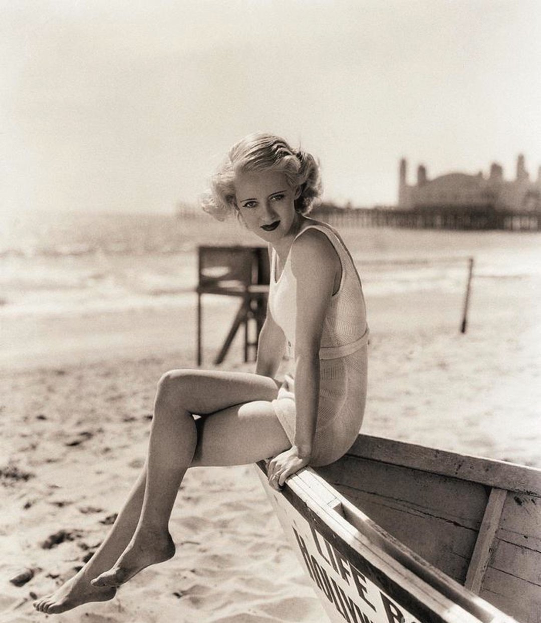 A woman in a vintage swimsuit sits on the edge of a wooden lifeboat on a sandy beach, with the ocean and a pier in the background. The photo is in black and white, evoking a classic, old Hollywood feel.