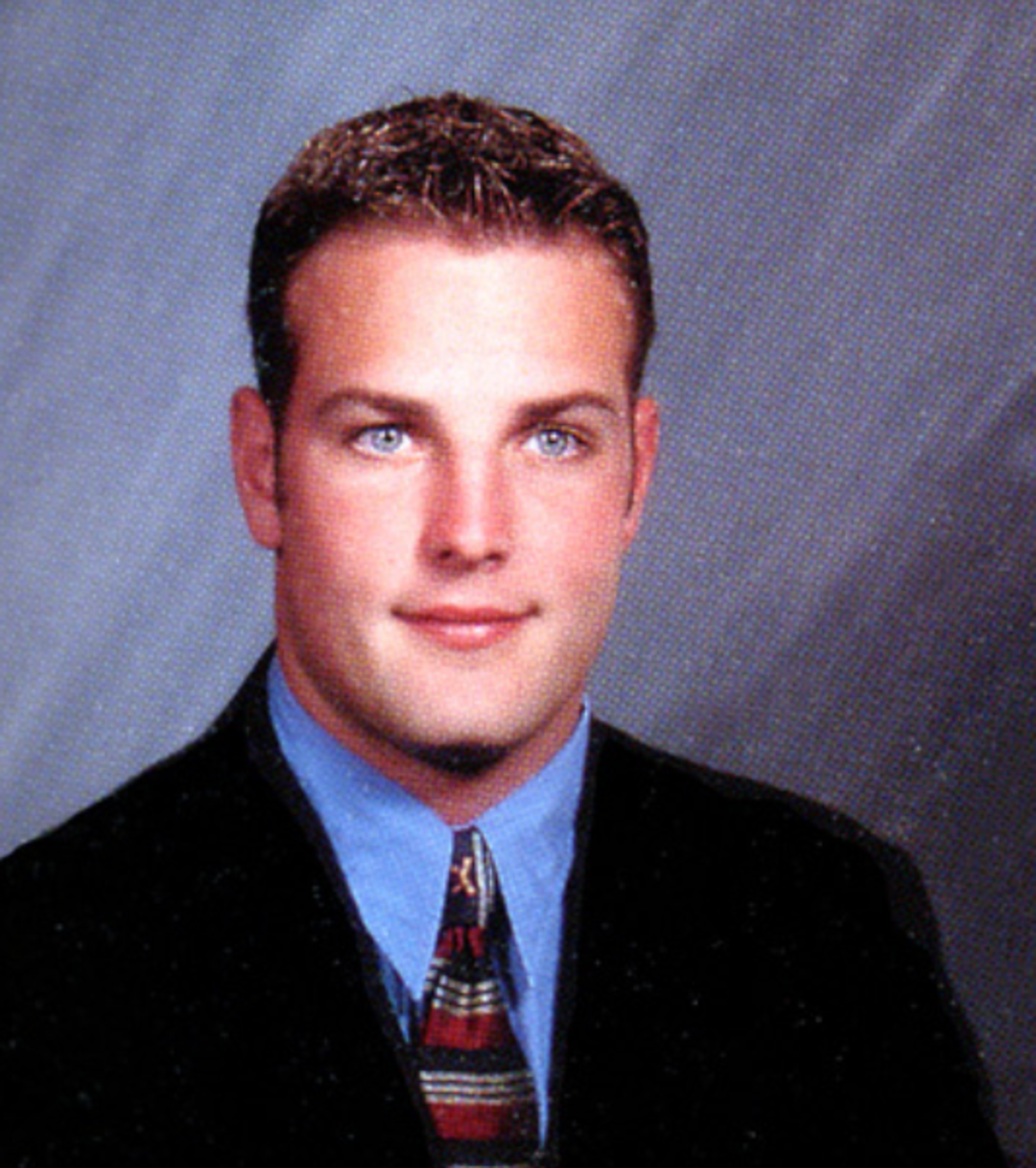 A young man in a suit, blue shirt, and patterned tie poses for a formal studio portrait against a gray background. He has short hair and a slight smile.