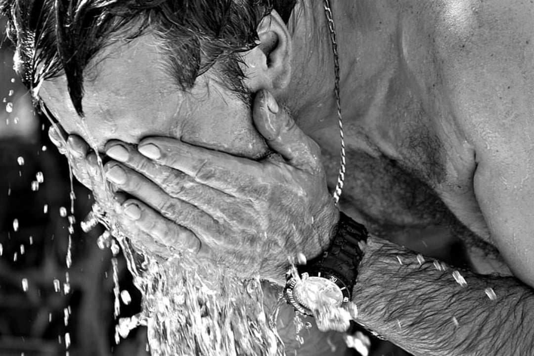 A shirtless man wearing a watch and necklace splashes water on his face, covering it with both hands. The image is in black and white.
