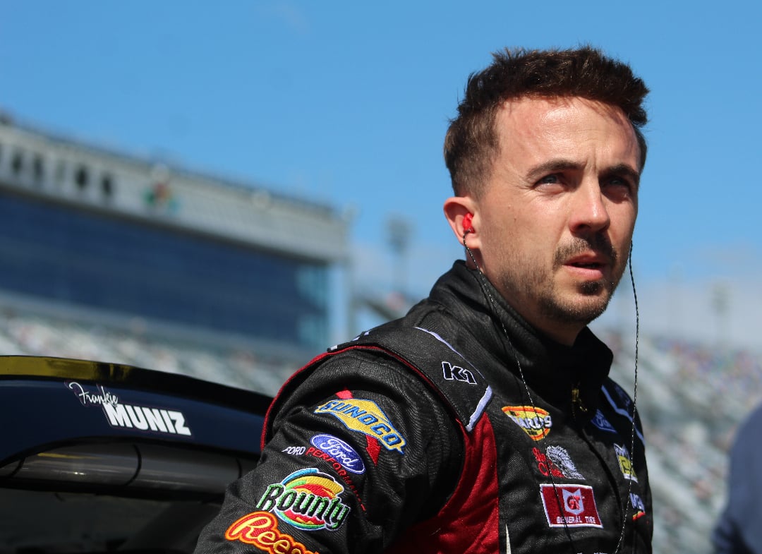 A race car driver in a black suit with sponsor logos stands near a car on a sunny day at a racetrack, looking into the distance with a focused expression.