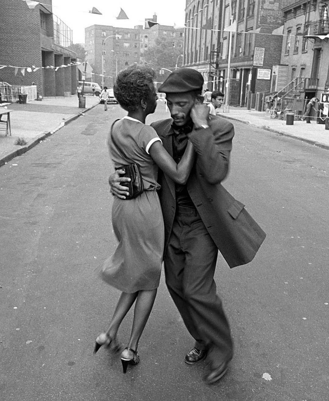 A man and woman dance closely together on an empty city street lined with buildings and decorations, capturing a joyful, lively moment in an urban neighborhood.