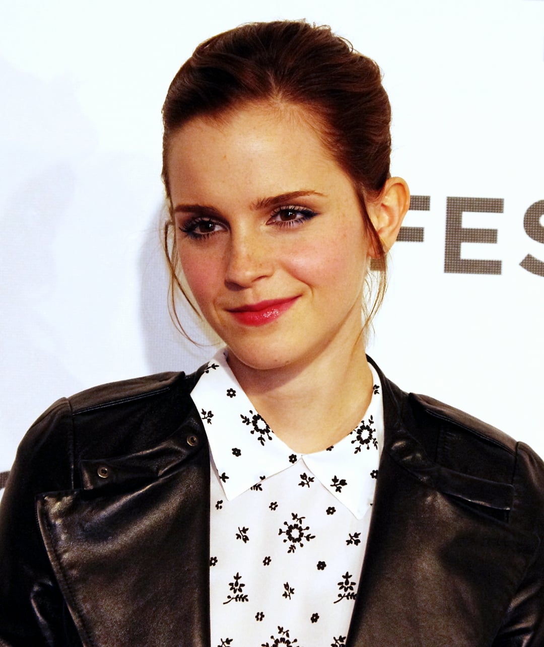 A woman with fair skin and brown hair tied back, wearing a black leather jacket over a white collared shirt with a black floral pattern, poses and smiles slightly in front of a light background.