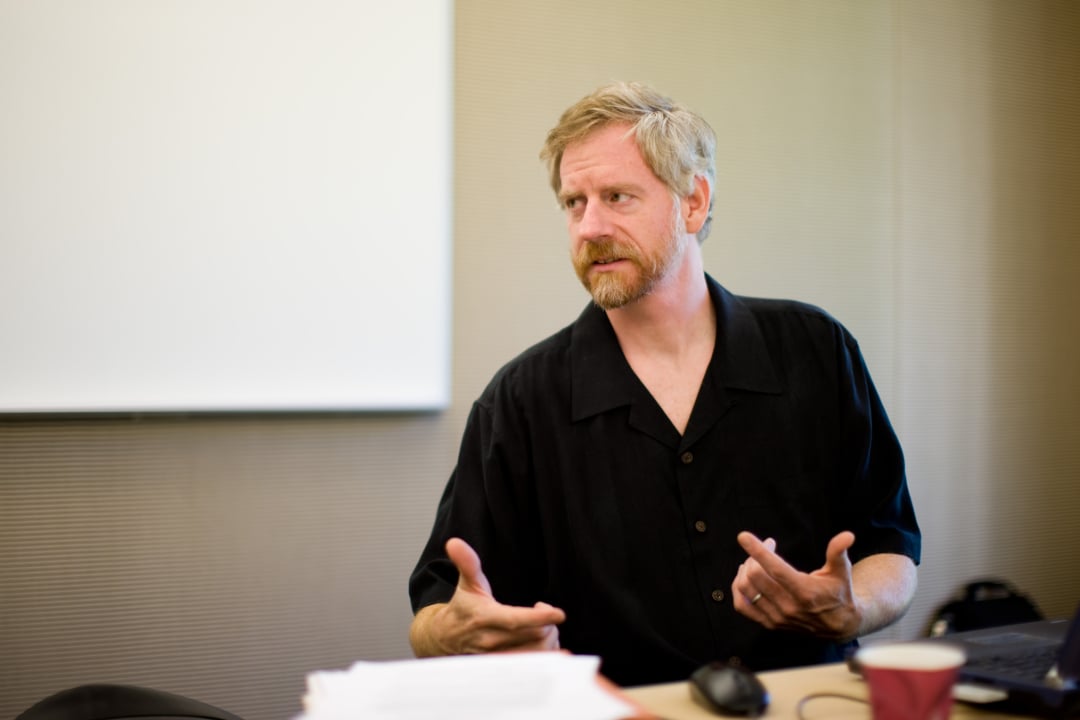 A man with light hair and a beard, wearing a black shirt, sits at a table gesturing with his hands. A stack of papers, a coffee cup, and a laptop are on the table in front of him. A blank screen is in the background.