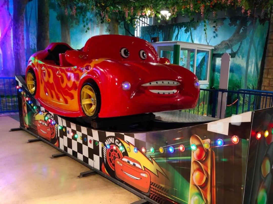 A red car ride for children, styled with cartoon eyes and a smile, sits on a platform decorated with colorful lights and racing graphics. The background features painted trees and a green booth.