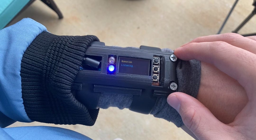 A person wears a homemade wrist device with buttons, a small screen displaying "Question Listening...", and a glowing blue light. The device is attached over a sleeve with screws visible on its case.