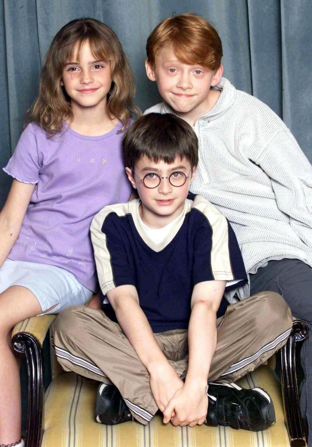 Three children pose together on a striped sofa in front of a grey curtain. The girl on the left wears a purple shirt, the boy behind wears a striped hoodie, and the boy in front wears glasses and a dark T-shirt.