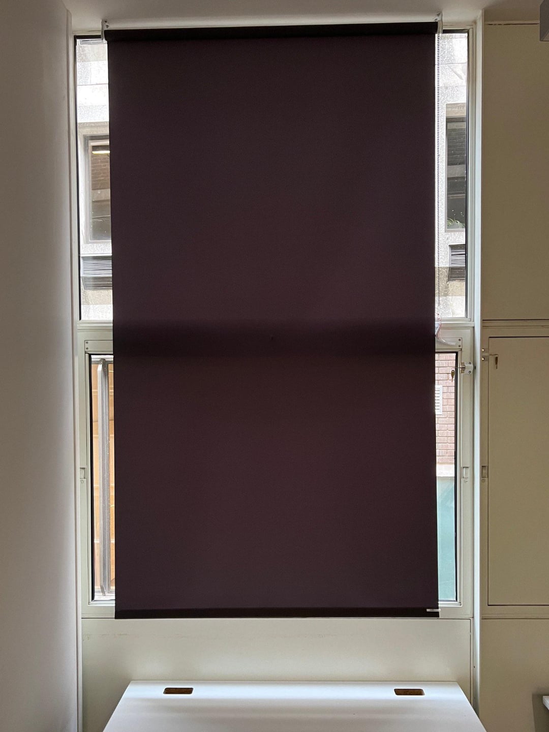 A dark roller blind is partially pulled down over a window, covering most of the glass. The window frame and part of a white wall and cabinet are visible around the edges.