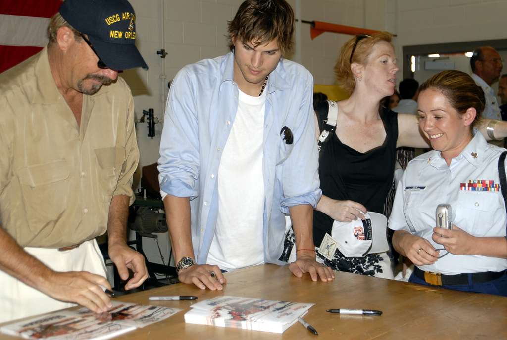 Three people, including a man in a "USCG Air New Orleans" cap, a casually dressed man, and a woman in a military uniform, sign autographs at a table while others interact nearby in what appears to be a public event indoors.