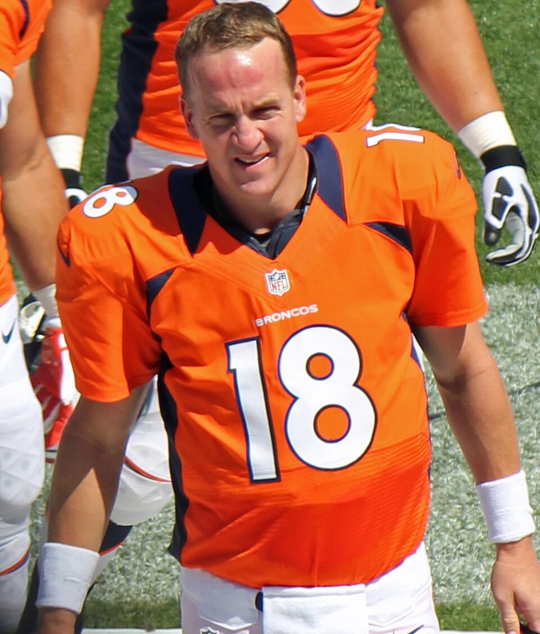 A football player in an orange Denver Broncos jersey with the number 18 stands on the field, squinting and smiling in the sunlight.