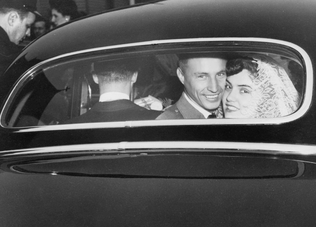 A smiling bride and groom sit in the backseat of a car, with the bride wearing a lace veil. The photo is black and white, capturing a joyful moment after their wedding ceremony.