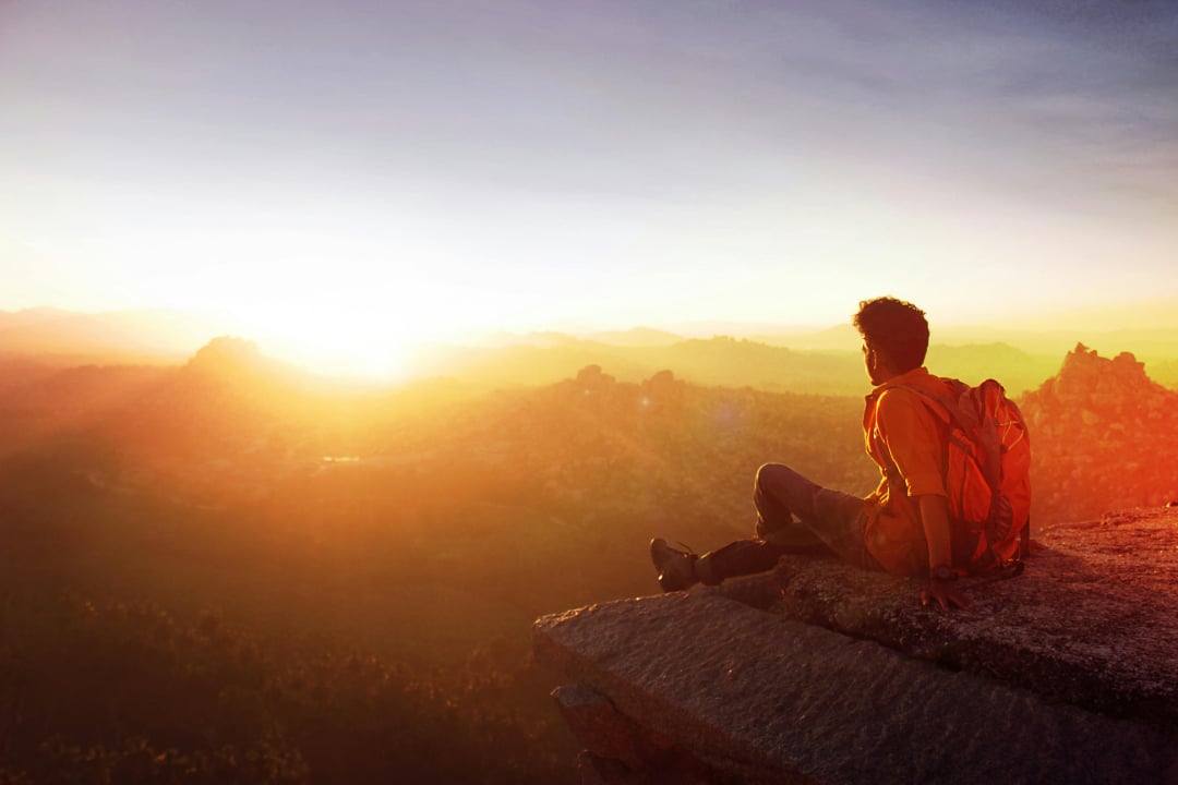 A person with a backpack sits on a rocky cliff edge, gazing at a glowing sunrise over a vast, mountainous landscape. The scene is bathed in warm sunlight.