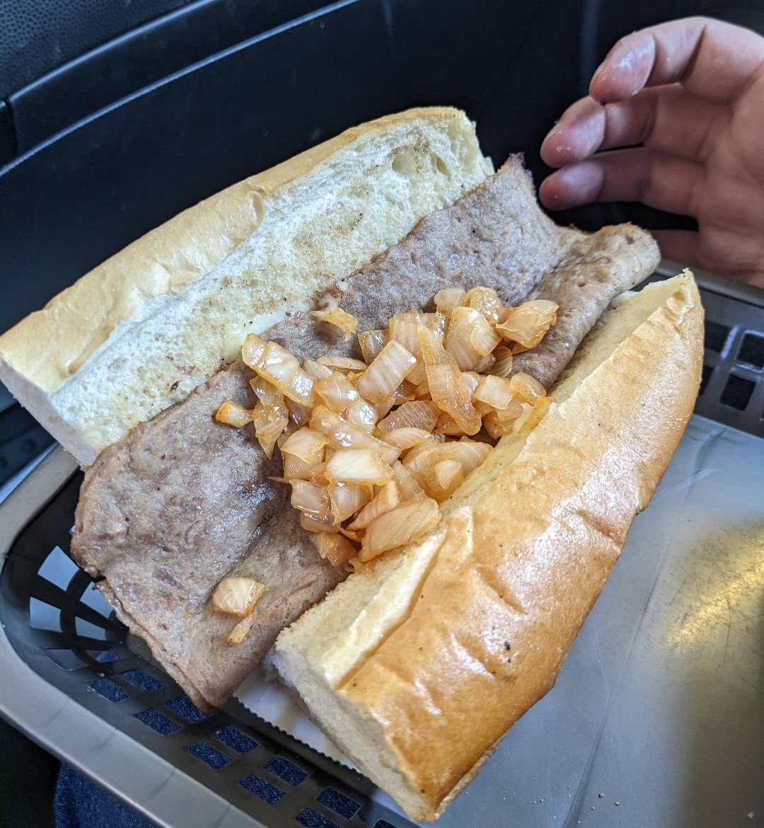 A sandwich roll filled with a thin slice of meat and chopped cooked onions, served in a plastic basket. A person’s hand is visible near the food.